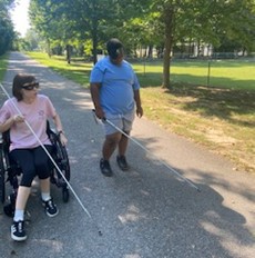 Mentor in wheelchair and white cane travels along side student with learning shades and white cane on park path